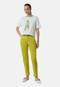 Witte T-shirt met een grafiek van een zittende figuur, gecombineerd met felle gele broek en witte schoenen, benadrukt door een bruine riem.