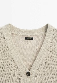 Cardigan beige en maille ajourée avec col en V, fermeture par un bouton noir unique et étiquette de marque noire visible à l’intérieur du col.