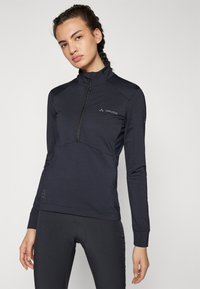 Haut mi-zippé bleu marine foncé en matériau léger, avec des manches longues et une coupe ajustée, arborant un branding discret sur la poitrine et la manche.
