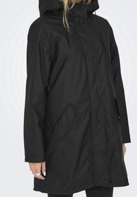 Femme portant un imperméable noir à capuche avec des boutons-pression à l'avant et des poches à rabat inclinées, debout devant un fond clair uni.