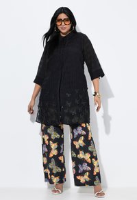 Tunika nera semi-trasparente con ricami a farfalla, abbinata a pantaloni a gamba larga con stampa di farfalle colorate. Sandali bianchi completano il look.