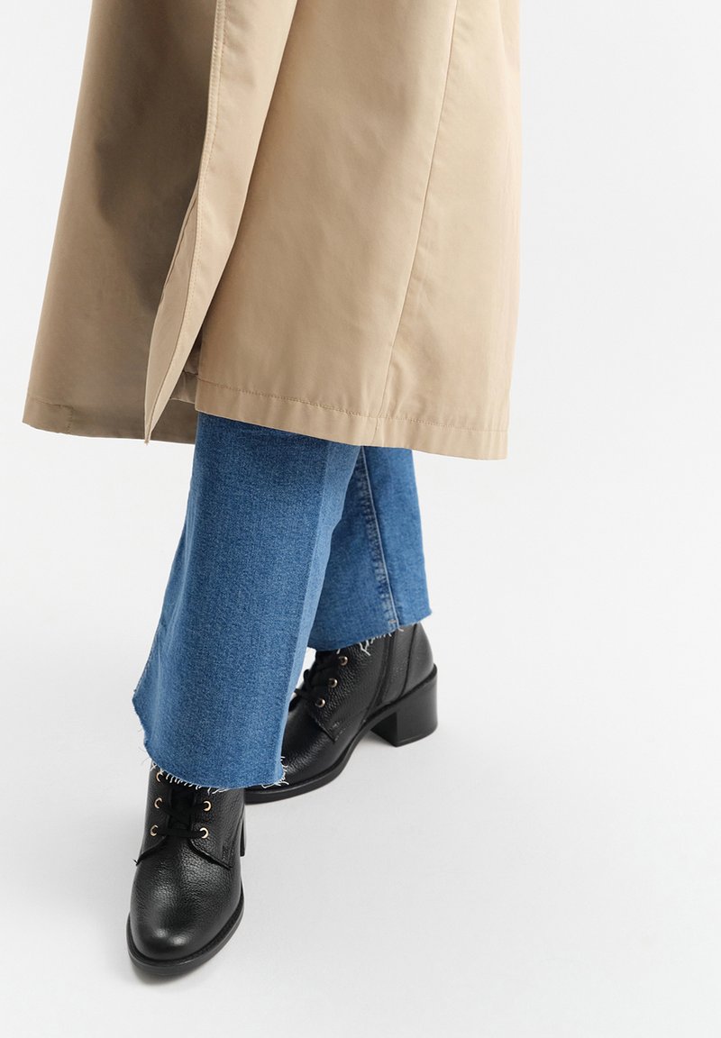 Czarne skórzane botki za kostkę z grubym obcasem, w połączeniu z postrzępionymi niebieskimi dżinsami i beżowym długim płaszczem.