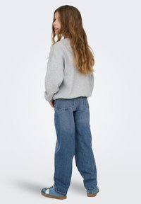 Szürke pulóver, bő kék farmer és kék tornacipők. Hosszú, hullámos barna haj. A ruha lazán szabott, kényelmes, hétköznapi viselet.