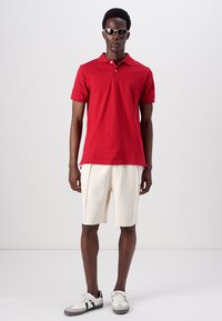 Polo shirt en coton rouge avec un petit logo, associé à un short crème et des baskets grises. Modèle portant des lunettes de soleil, se tenant devant un fond blanc.