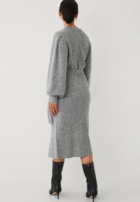 Grijze gebreide midi-jurk met lange, opgeblazen mouwen en een strikceintuur in de taille. Geribbelde textuur en zijsplit. Gemaakt met zwarte kniehoge laarzen.