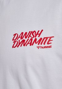 Rode tekst "DANISH DYNAMITE" en rood hummelbijlogo op witte stof, waarschijnlijk een shirt of een sweater.
