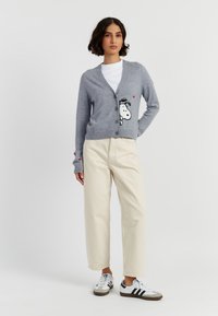 Lichtgrijze gebreide cardigan met een knoopsluiting, voorzien van een Snoopy-afbeelding op de voorkant, gecombineerd met crèmekleurige broek en casual sneakers.