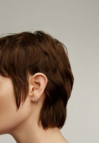 Pendiente de aro de plata con pequeños acentos circulares, usado en la oreja. Cabello corto y texturizado de color marrón que enmarca parcialmente la oreja.