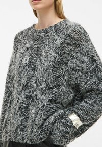 Sweater de gran tamaño en gris y negro tejido, que presenta un patrón de trenzas. Incluye un acento metálico en el puño y un material texturizado y esponjoso.