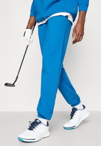 Zilas sporta bikses ar ribotām piedurknēm, kombinētas ar baltām golfa kurpēm ar tumši ziliem akcentiem. Modelis tur golfa nūju, kamēr iet.