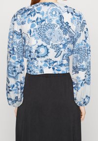 Blusa leggera azzurro chiaro con stampa floreale, maniche lunghe a palloncino e design cropped, caratterizzata da motivi intricati su uno sfondo bianco.