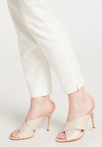 Beige hælede mules med et krydsdesign, lavet af glat læder, med spids tå og stilettohæl. Båret med hvide bukser.