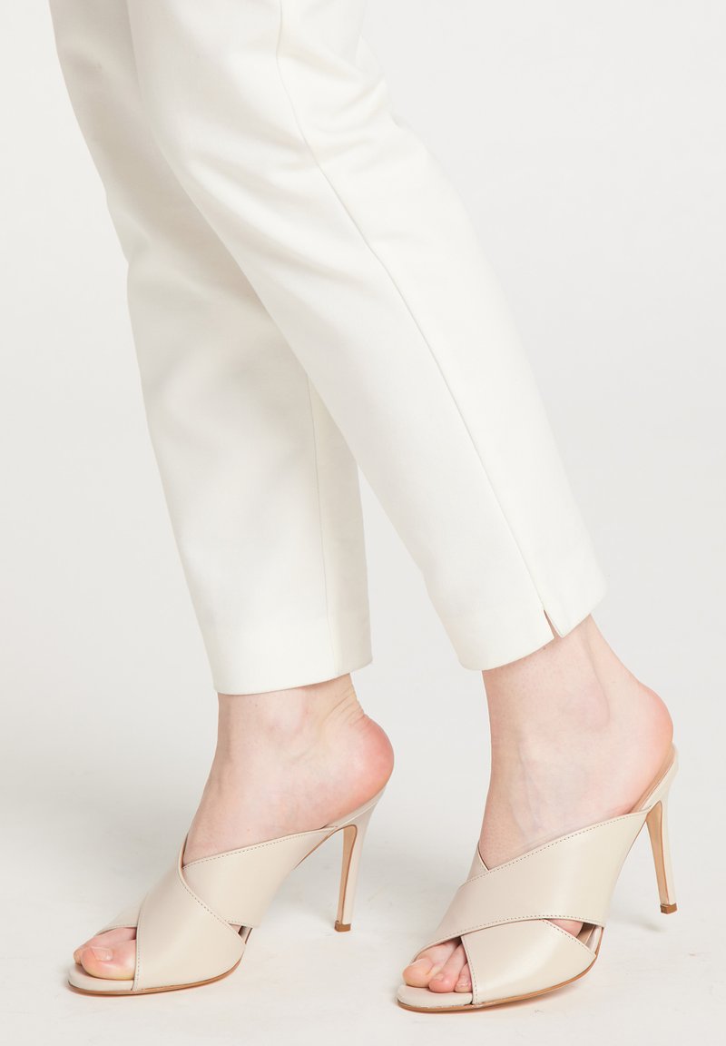 Beige hælede mules med et krydsdesign, lavet af glat læder, med spids tå og stilettohæl. Båret med hvide bukser.