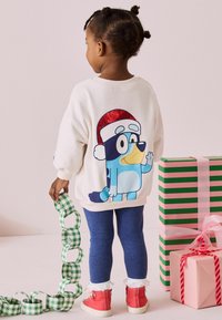 Biała bluza z kapturem z niebieską postacią z kreskówki i czerwonym błyszczącym kapeluszem Świętego Mikołaja. Niebieskie legginsy i czerwone buty z koronkowym wykończeniem. Zielony łańcuch dekoracyjny z papieru w kratkę.