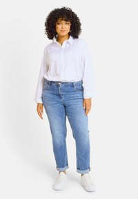 Chemise blanche à manches longues, associée à un jean bleu clair à ourlets retroussés et des baskets blanches, mettant en avant un look décontracté.