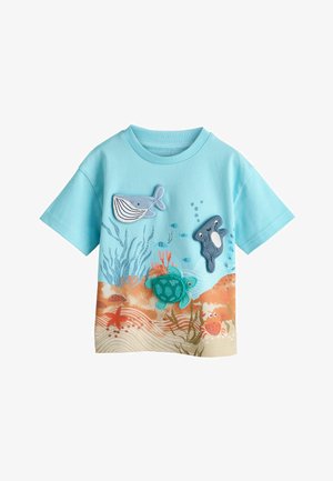 Lyseblå T-shirt med korte ærmer med applikationer af vanddyr: hval, skildpadde, fisk og krabbe på en sandet havbundsbund.