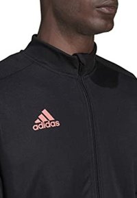 Giacca nera con zip realizzata in tessuto leggero, presenta un colletto alto e un logo Adidas rosa sulla parte sinistra del petto.