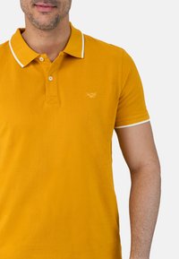 Una camiseta polo amarilla con un tejido texturizado, un cuello clásico, detalles en blanco en el cuello y las mangas, y un pequeño logo bordado en el pecho.
