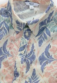 Chemisier à manches courtes en tissu léger, avec un motif floral bleu et rose, des découpes et des boutons en perles.