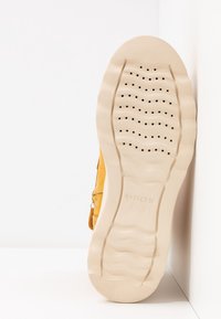 Semelle de chaussure en caoutchouc beige avec motifs ondulés et perforations pour la respirabilité, partie supérieure jaune partiellement visible, sur fond blanc.
