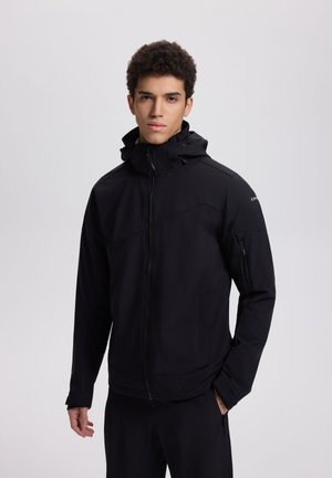 Junger Mann mit lockigem Haar, der eine schwarze Kapuzenjacke mit Reißverschluss und schwarze Hosen trägt und vor einem schlichten hellgrauen Hintergrund steht.