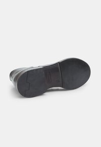 Suela de zapato de cuero negro con un acabado texturizado, forma ligeramente curvada y un tacón liso, que presenta un branding sutil y acentos remachados.