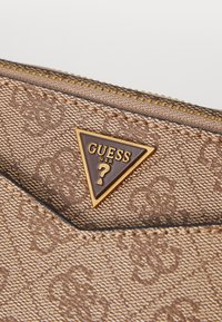Ruskean kuviollinen kangaslompakko, jossa on kultainen vetoketjutakki ja kolmiomainen metallilogo, jossa lukee "GUESS USA".