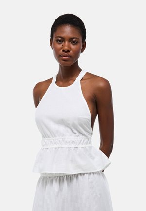 Femme aux cheveux courts portant un top péplum blanc sans manches et une jupe assortie sur un fond gris clair uni.