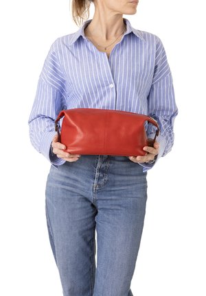 Donna con camicia a righe blu e jeans che tiene una pochette rossa in pelle con entrambe le mani all'altezza della vita.