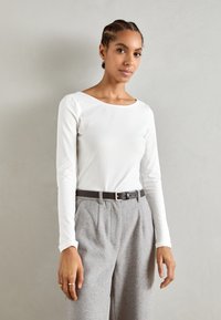 White long-sleeve top with a bateau neckline, paired with gray high-waisted trousers and a black belt. Smooth fabric, relaxed fit.