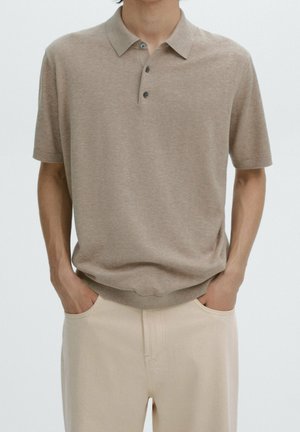 Mann trägt ein beigefarbenes Poloshirt mit kurzen Ärmeln und drei Knöpfen, die Hände in den Taschen einer cremefarbenen Hose, vor einem einfarbigen Hintergrund.