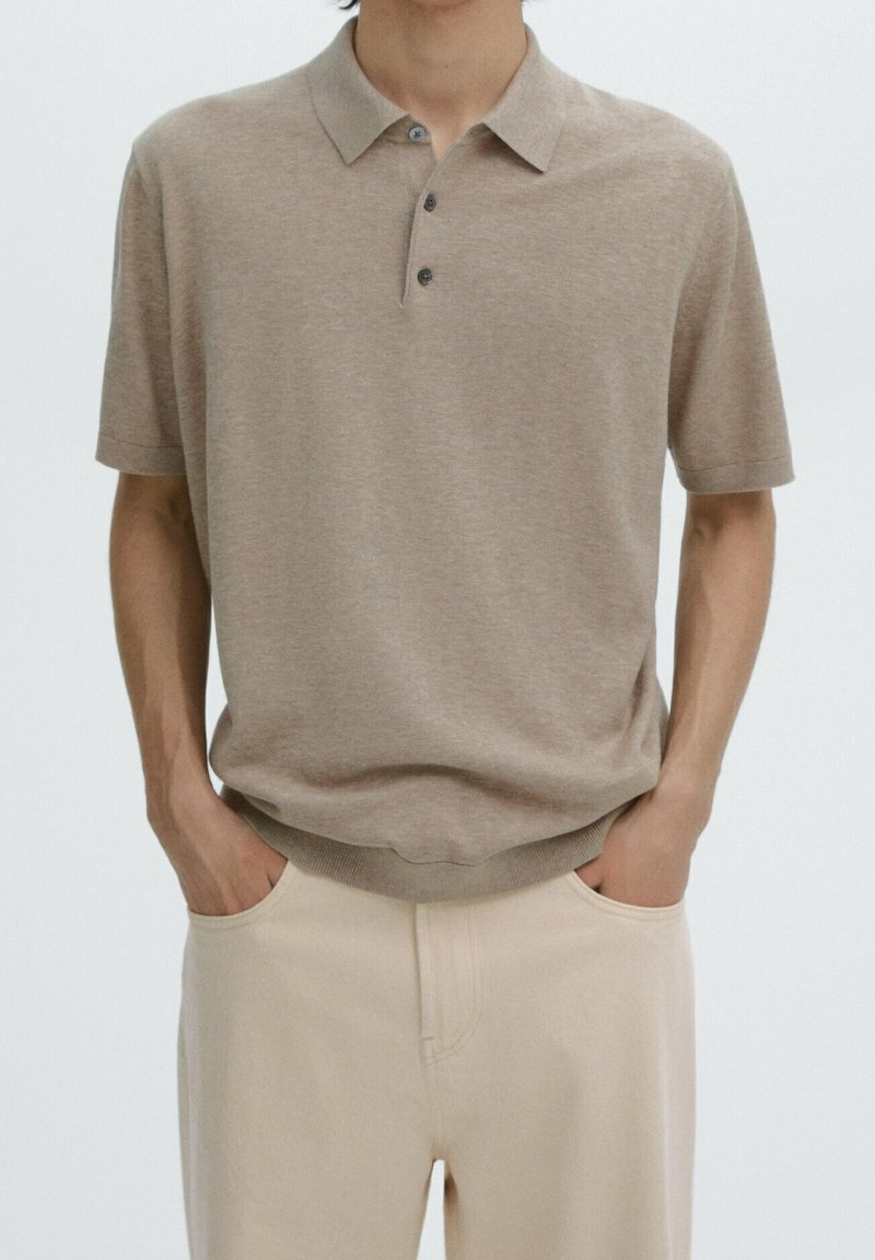 Homme portant un polo beige à manches courtes avec trois boutons, les mains dans les poches d'un pantalon de couleur crème, sur un fond uni.