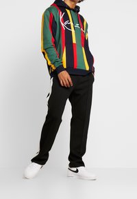 Persona vestida con una sudadera a rayas de colores, pantalones negros con franjas laterales blancas y zapatillas Nike blancas, de pie contra un fondo liso.