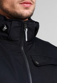 Námořnická modrá bunda vyrobená z odolné tkaniny, s vysokým límcem, plným zipem a matným stříbrným logem na kapsičce na hrudi.