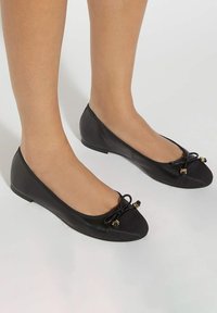 Black leather ballet flats with a black fabric cap toe, bow detail, and gold accents. Slight heel and smooth texture.