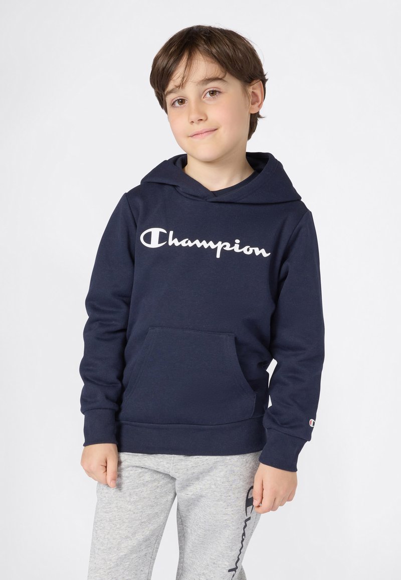 Jongen met kort bruin haar, gekleed in een marineblauwe Champion hoodie en lichtgrijze Champion joggingbroek, staande tegen een witte achtergrond.