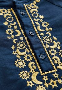 Tissu bleu foncé avec des motifs célestes brodés jaunes, incluant des étoiles et des croissants de lune autour d'une patte de boutonnage.