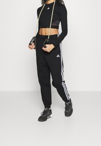 Svart långärmad tröja med Adidas-logga, svarta träningsbyxor med vita sidostreck och svarta sneakers, håller i träningsresistansband.