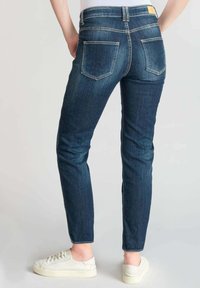 Jean en denim bleu foncé avec une coupe ajustée, présentant deux poches arrière et des coutures claires. Associé à des baskets blanches.