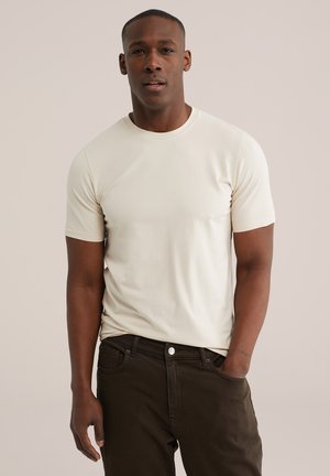 Man draagt een effen off-white korte mouwen T-shirt en donkerbruine broek, staand met één hand in de zak tegen een effen achtergrond.