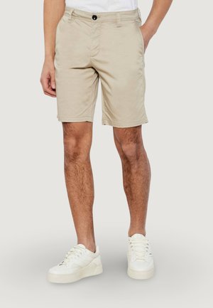 Uomo che indossa pantaloncini beige fino al ginocchio, scarpe da ginnastica bianche e una camicia bianca, in piedi con una mano in tasca contro uno sfondo semplice.