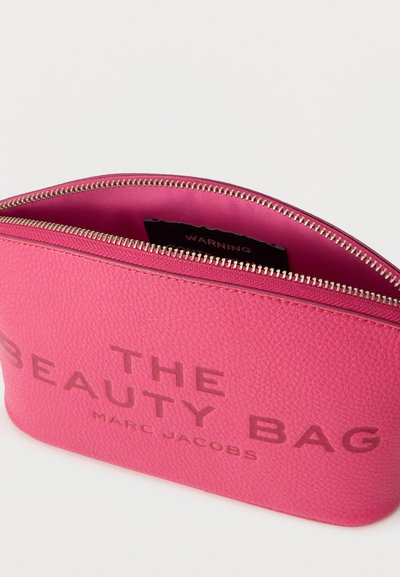 Φωτεινό ροζ υφασμάτινο τσαντάκι με φερμουάρ και την επιγραφή «THE BEAUTY BAG MARC JACOBS» στην πρόσοψη, μερικώς ανοιχτό για να φαίνεται το εσωτερικό ύφασμα και η ετικέτα.