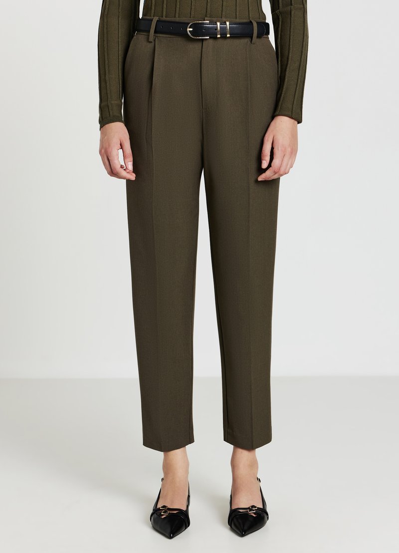 Personne portant un pantalon taillé vert olive avec une ceinture noire et des chaussures noires pointues, se tenant contre un fond clair.