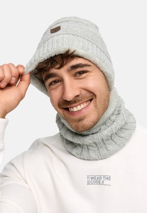 Bonnet en tricot gris avec un motif texturé et un patch logo, accompagné d'un cache-cou assorti. Tissu doux à côtes ; design confortable.