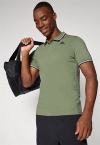 Olivenfarget polo-skjorte med korte ermer, ribbestrikket krage med blå aksenter og en liten svart logo. Modellen holder en svart duffelbag.