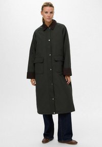 Cappotto lungo verde scuro con colletto marrone, chiusure a scatto frontali e due tasche laterali; il tessuto sembra liscio, maniche con texture.
