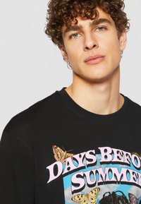 Black cotton t-shirt featuring a graphic print with the text "DAYS BEFORE SUMMER" in pink, surrounded by butterfly illustrations.