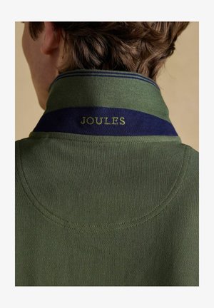 Person iført grøn polo med navyblå kant på kraven, der viser "JOULES" broderet på bagsiden af kraven.
