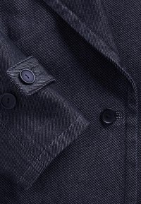 Sötétkék dzseki halszálka mintával, fekete gombokkal és testreszabott gallérral. Közeli kép az ujja és a gombok részleteiről.