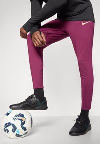Calças de treino Nike em marrom profundo, ajuste slim, feitas de tecido elástico. Ténis pretos com detalhes em azul-turquesa. Bola de futebol branca debaixo do pé.